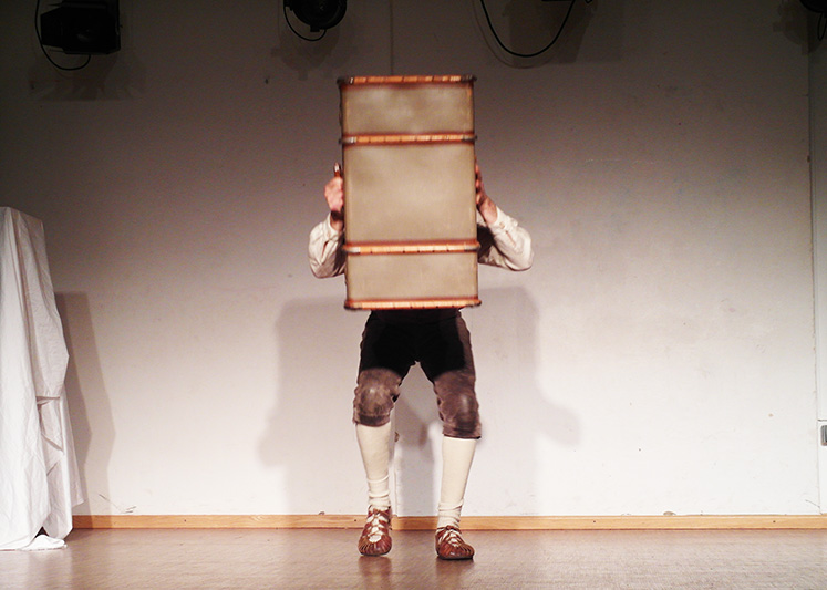 Monolog: Mann in Lederhose hält sich hinter Reisekoffer vor Kopf und Oberkörper